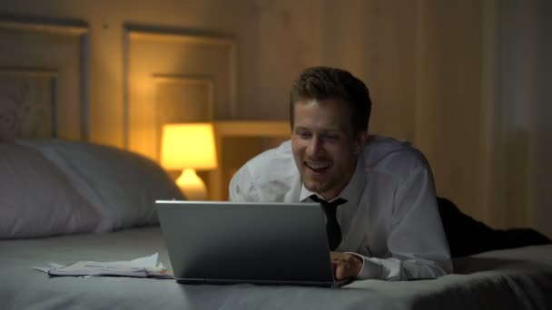 Smiling male in white shirt on bed smiling and chatting on laptop, office affair - Кадры, видео