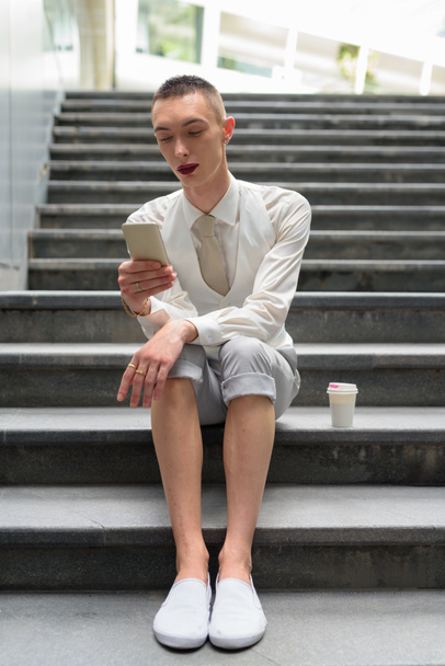 Νεαρός ανδρόγυνο ομοφυλοφιλικές επιχειρηματίας Lgtb κάθεται σε σκάλες, κατά τη χρήση του τηλεφώνου - Φωτογραφία, εικόνα