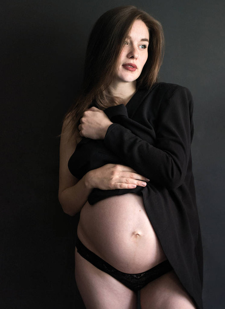 foto portret van een mooie zwangere meisje in een zwarte jas en slipje op een donkere achtergrond - Foto, afbeelding