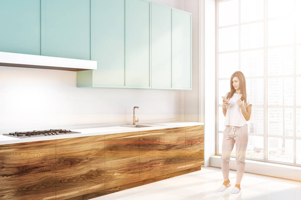 Woman standing in modern kitchen interior with white walls and floor, wooden countertops with built in appliances and blue cupboards. Toned image - Foto, afbeelding