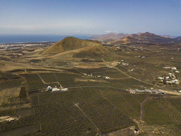 Vista aérea dos cultivos de vinho nos solos vulcânicos da ilha de Lanzarote, planícies e colinas no interior, vulcões no horizonte. Ilhas Canárias, Espanha. Produção de vinho
 - Foto, Imagem
