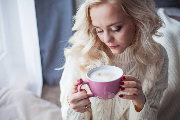 Cozy home portrait of a young beautiful woman. A beautiful blonde girl in a knitted dress is sitting by the window drinking a cappuccino and reading a book. Autumn decor. Cozy. Winter. Autumn. - Valokuva, kuva