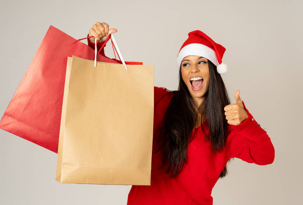 Young woman ready for christmas with paper shopping bags in red and Santa claus hat isolated grey background in being ready for christmas shopping gifts celebration consumerism and sales concept. - Photo, image