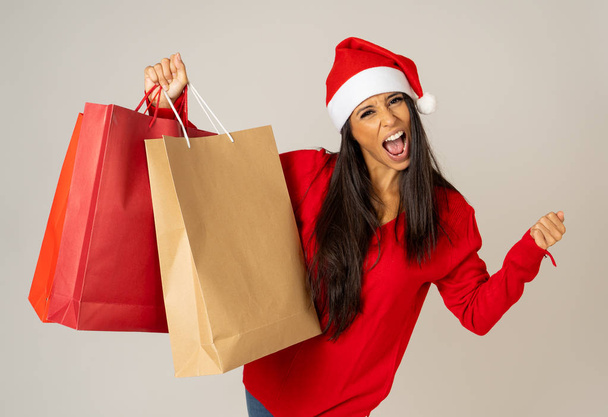 Young woman ready for christmas with paper shopping bags in red and Santa claus hat isolated grey background in being ready for christmas shopping gifts celebration consumerism and sales concept. - Photo, Image