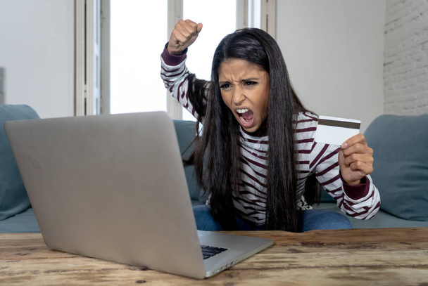 Attractive young latin woman desperate and furious with credit card and laptop siting on sofa home in credit card problems debts online payment online shopping online banking bad financial situation. - 写真・画像