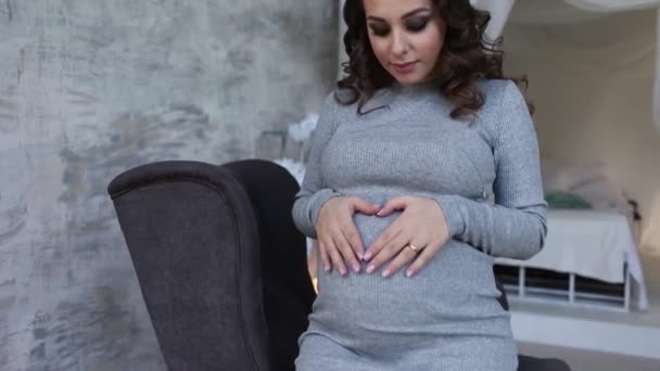 Beautiful pregnant woman sit on the armchair and put her hands to her belly. Woman dressed in elegant dress. Beautiful hairstyle and makeup - Кадри, відео