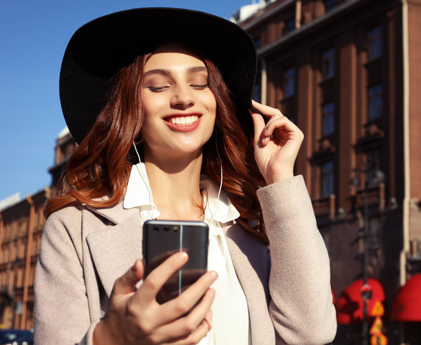 schönes junges Mädchen in lässigem Stil gekleidet - Mantel und Hut, Musik hören und SMS auf Smartphone-Gerät tippen. - Foto, Bild