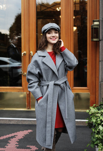 young stylish pretty woman wearing red dress, grey coat and hat posing in the city streets. - Foto, Bild