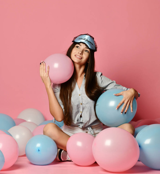 Cute young woman in pyjamas and sleepmask having fun with helium balloons. portrait of pleasant birthday girl on pink background. - Foto, imagen