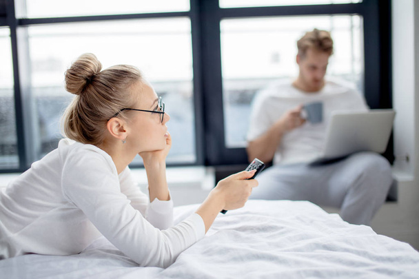 serioous fair-haired girl is watching TV while her husband surfing the net - Foto, Bild