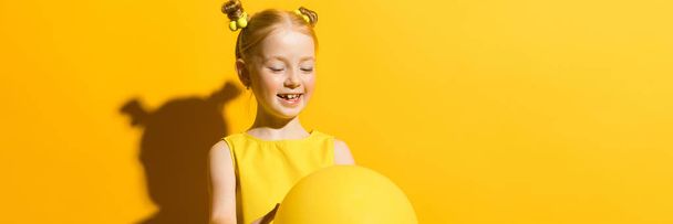 Fille aux cheveux roux sur fond jaune. La fille tient dans ses mains et regarde le ballon jaune
. - Photo, image