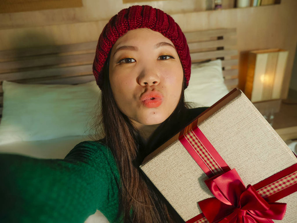 молодая красивая и счастливая азиатская американская девушка делает селфи фото с мобильного телефона, держа рождественский подарок коробка улыбаясь веселый и взволнованный зимой сияющий дома празднует Рождество праздники
 - Фото, изображение