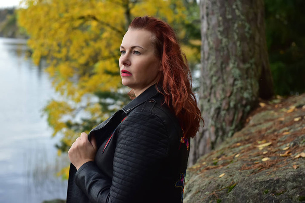red-haired woman in a black leather jacket on an autumn background - Photo, Image