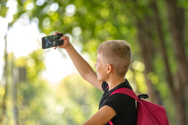 young happy preteen boy walking in warm sunny summer day in the park and taking selfie on smartpone - Фото, зображення