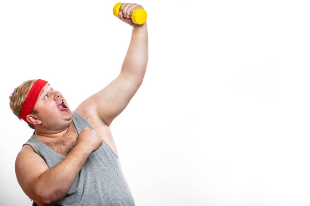 Fat funny man in red headband shows his muscles with dummbell and emotions - Foto, Bild