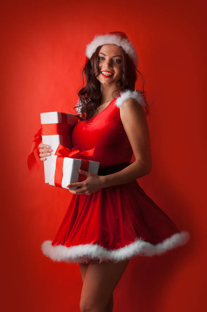 Beautiful young woman in Santa dress and hat celebrating Christmas holding gift boxes - Foto, afbeelding