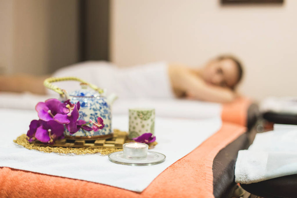 Young beautiful girl in spa salon. The girl lies on the couch for a massage after a rejuvenation procedure. a composition in Thai style with an orchid and a teapot with green tea - Fotoğraf, Görsel