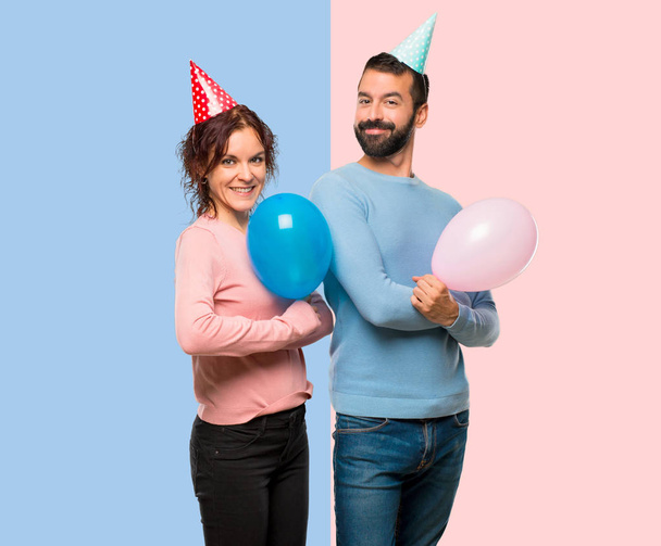 couple with balloons and birthday hats keeping the arms crossed in lateral position while smiling on pink and blue background - Photo, Image