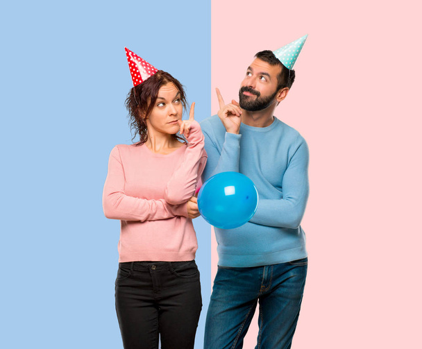 couple with balloons and birthday hats pointing with the finger a great idea and looking up on pink and blue background - Photo, Image
