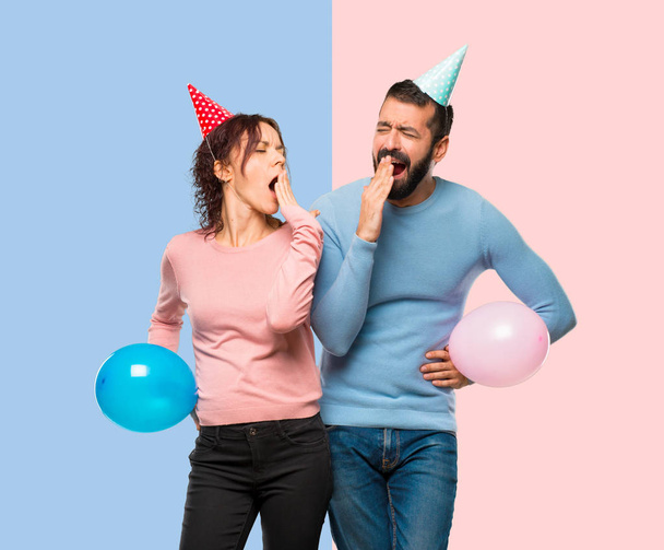 couple with balloons and birthday hats yawning and covering mouth with hand. Sleepy expression on pink and blue background - Photo, Image