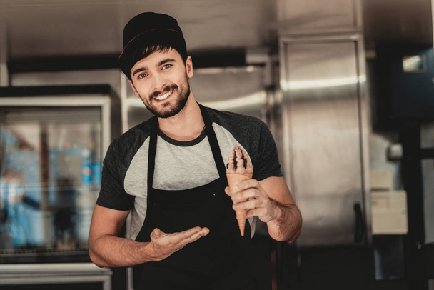 Young Bearded Man in Apron Standing in Food Truck. Ice Cream in Hand. Street Food Concept. Food in Town. Selling Snacks. Guy in Cap. Black Arpon. Working Outdoor. Man in T-shirt. Standing Guy. - Fotografie, Obrázek