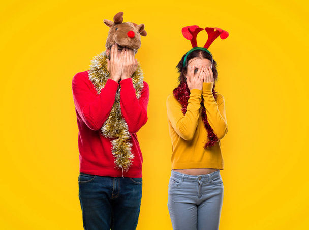 Couple dressed up for the christmas holidays covering eyes by hands and looking through the fingers on yellow background - Foto, Bild