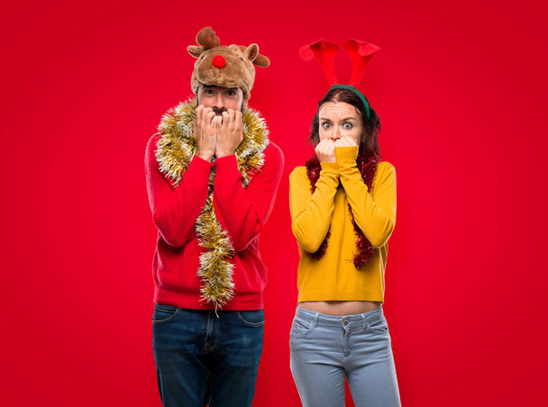Casal vestido para as férias de Natal é um pouco nervoso e assustado colocando as mãos na boca em fundo vermelho isolado
 - Foto, Imagem