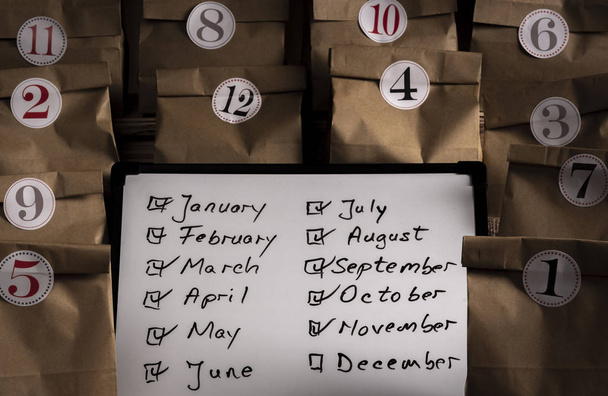 Writing board with the list of the months of the year and paper bags numbered from 1 to 12 with stickers. Traditional advent calendar gifting in small paper bags - Photo, Image