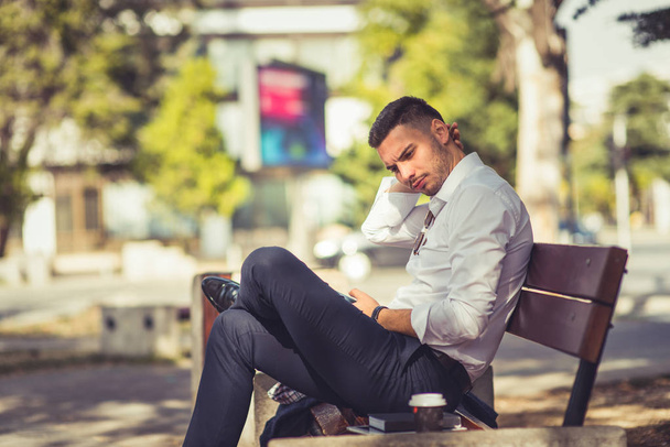 Современный бизнесмен сидит в парке на скамейке со скрещенными ногами, и он выглядит любопытным и серьезным
 - Фото, изображение