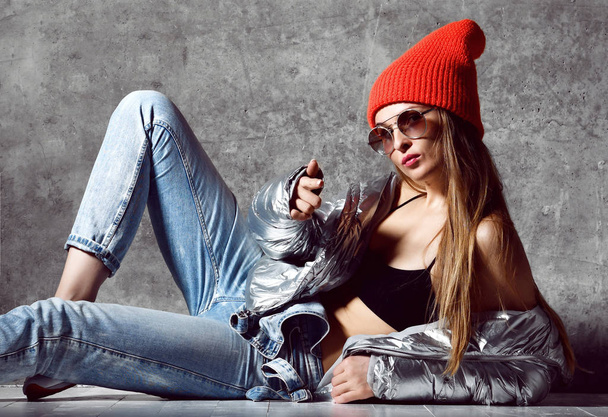 Young sport woman after workout exercise sitting point finger in silver sexy thin down puffer jacket blue jeans red hat  - Fotoğraf, Görsel