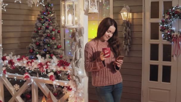 Γυναίκα, που χαμογελά ποτά τσαγιού χρήση τηλέφωνο στην βεράντα με το χριστουγεννιάτικο δέντρο στο παρασκήνιο - Πλάνα, βίντεο