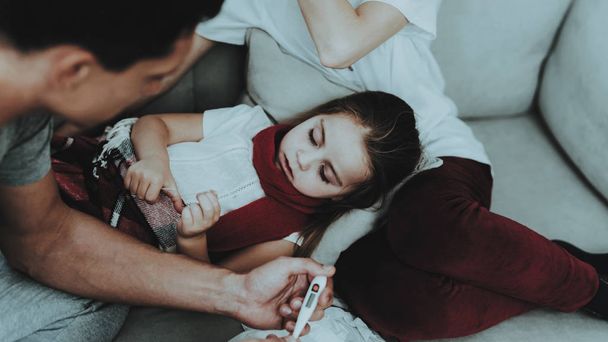 Little Girl in Red Scarf with Cold with Family. Sick Young Girl. White Sofa in Room. Father and Mother. Disease Concept. Healthcare and Healthy Lifestyle Concept. Take Care of Daughter. - Zdjęcie, obraz