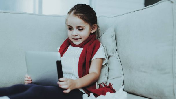 Little Girl in Red Scarf with Cold Using Tablet. Sick Young Girl. White Sofa in Room. Unhappy Child. Disease Concept. Healthcare and Healthy Lifestyle Concept. Little Girl with Tablet PC. - Fotografie, Obrázek