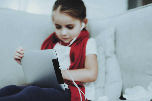 Little Girl in Red Scarf with Cold Using Tablet. Sick Young Girl. White Sofa in Room. Unhappy Child. Disease Concept. Healthcare and Healthy Lifestyle Concept. Little Girl with Tablet PC. - Photo, image