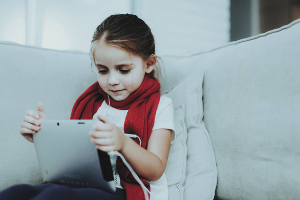 Little Girl in Red Scarf with Cold Using Tablet. Sick Young Girl. White Sofa in Room. Unhappy Child. Disease Concept. Healthcare and Healthy Lifestyle Concept. Little Girl with Tablet PC. - Foto, Bild