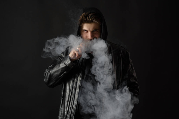 Mann mit dampfendem Mod, der Dampf vor schwarzem Studiohintergrund ausatmet. bärtiger Typ, der E-Zigarette raucht, um mit dem Tabak aufzuhören. Dampf und alternatives nikotinfreies Raucherkonzept - Foto, Bild