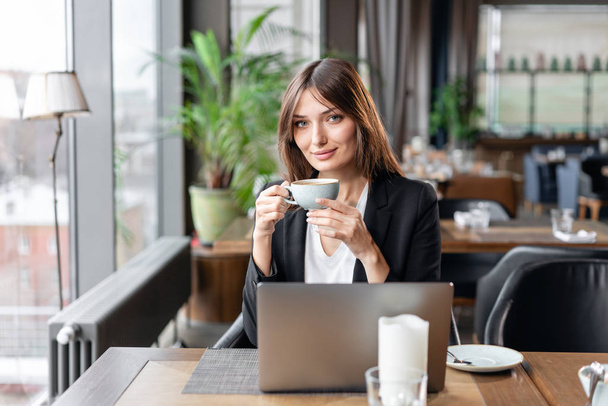 Jonge vrouw drinken koffie. Vrouwelijke bedrijf cup. Zittend in de coffeeshop op houten tafel. Is grijs aluminium laptop op tafel. Meisje bloggen, surfen op internet, chatten - Foto, afbeelding