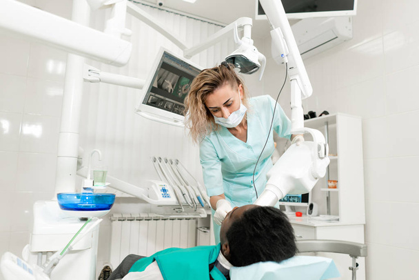Vrouw tandarts met behulp van x ray machine, patiënt liggend op de stoel in de tandheelkunde. Jonge African American man met slechte tanden. Geneeskunde, gezondheid, stomatologie concept. - Foto, afbeelding