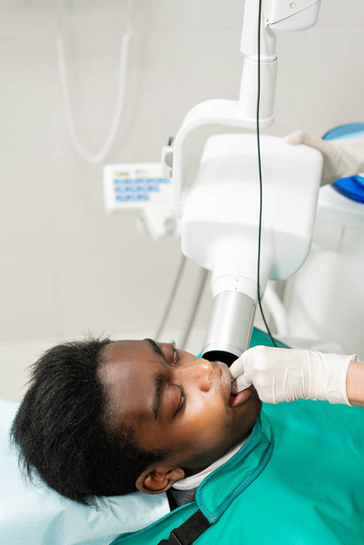 Mujer dentista usando máquina de rayos X, paciente acostada en silla en odontología. Joven hombre afroamericano con mal diente. Medicina, salud, concepto de estomatología
. - Foto, Imagen
