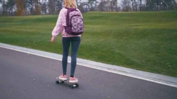 Young pretty beautiful blond hipster woman in pink windstopper having fun riding skateboard longboard downhill on beautiful road in slow motion - Imágenes, Vídeo