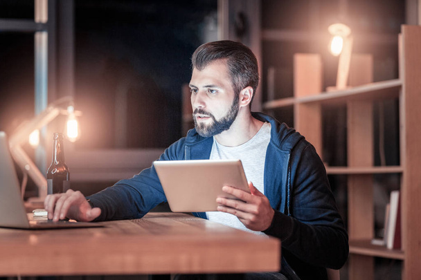 Let me check. Concentrated brunette man holding tablet in left hand and looking at computer while being at workplace - Foto, Bild