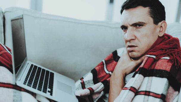 Man with Cold Sitting on Sofa in Checkered Blanket. Man at Home. Sick Young Man. White Sofa in Room. Unhappy Guy. Disease Concept. Healthcare and Healthy Lifestyle Concept. Using Laptop. - Foto, immagini