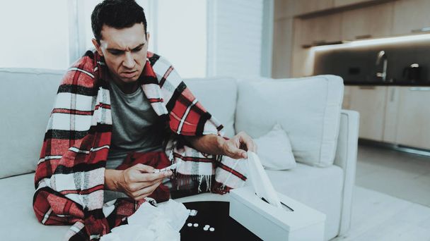 Man with Cold Sitting on Sofa in Checkered Blanket. Man at Home. Sick Young Man. White Sofa in Room. Unhappy Guy. Disease Concept. Healthcare and Healthy Lifestyle Concept. Tacking Pills. - Photo, image
