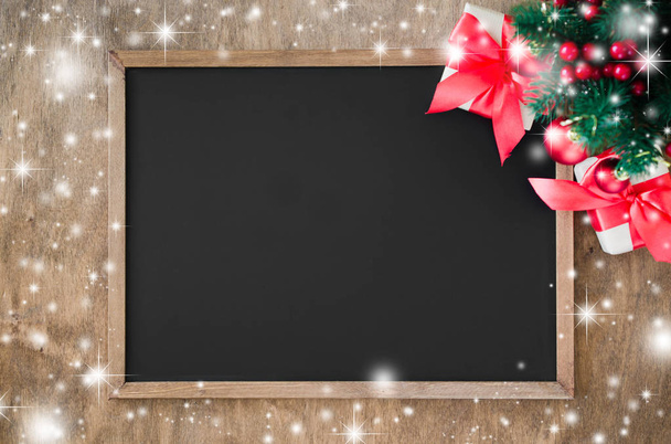 Empty blackboard with christmas decoration, red gift box, fir branches. Chalk board and Christmas trinkets. Xmas background with copy space for inscription, top view. Snow effect. - Photo, Image