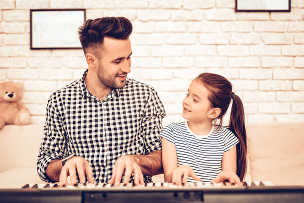 Man and Girl Play Piano. Play Piano Together. Father and Daughter on Sofa. Spend Time Together. Father's day. Man Play with Girl. White Interior. Girl wiht Man on Sofa. Play Piano and Sing. - Photo, image