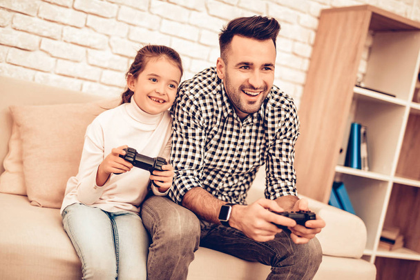 Joystick in Hand. Father and Daughter. Spend Time Together. Father's day. Man Playing with Girl. Video Game. White Interior. Sitting on Sofa. Girl with Joystick in Hands. Plaid Shirt. - Zdjęcie, obraz
