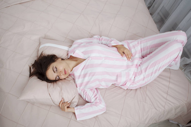 Fiatal aranyos sötét hajú nő, rózsaszín csíkos pizsamás feküdt egy ágyon, rózsaszín ágynemű és párnák. - Fotó, kép