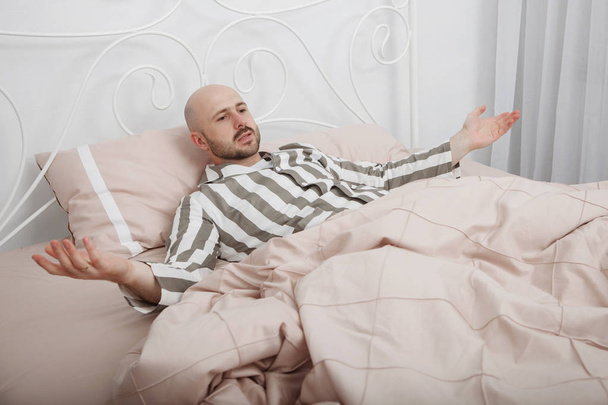Guapo calvo, barbudo hombre en pijama a rayas está acostado y pensando en la cama con almohadas y ropa de cama rosa
. - Foto, Imagen