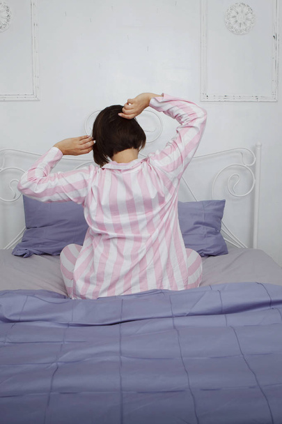 Fiatal aranyos sötét hajú nő rózsaszín pizsama ül az ágyon, kék ágynemű és párnák. Hátulnézet. - Fotó, kép