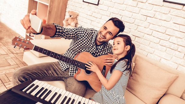 Man and Girl Play Piano. Play Guitar. Make Selfie Together. Piano and Guitar. Father and Daughter on Sofa. Spend Time Together. Father's day. Man Play with Girl. White Interior. Musical Instruments. - 写真・画像